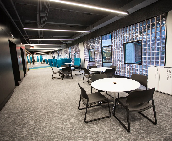 View down a wide corridor with elevators to the left and round tables surrounded by black chairs.